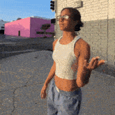 a woman wearing glasses and a white tank top is standing on a sidewalk in front of a pink building