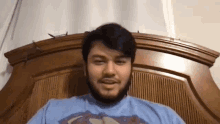 a man with a beard is sitting on a bed wearing a blue shirt with a superman logo on it .