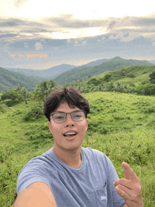 a young man wearing glasses takes a selfie in front of a grassy hillside