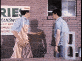 two men in aprons are standing in front of a brick building .