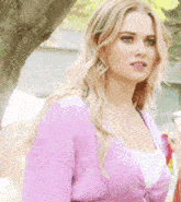 a blonde woman in a pink sweater is standing in front of a tree