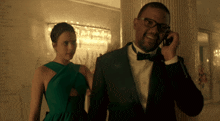 a man in a tuxedo talks on a cell phone while a woman in a green dress watches