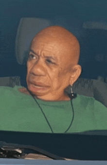 a bald man wearing a green shirt and ear buds looks out of a car window