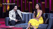 a man and a woman are sitting on a couch talking . the woman is wearing a yellow dress .