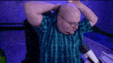 a bald man wearing glasses and a plaid shirt is sitting in front of a microphone and holding his head .