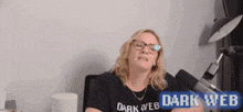 a woman wearing glasses and a black shirt that says dark web is sitting in front of a microphone