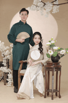 a man holding a fan stands next to a woman sitting in a chair holding a fan