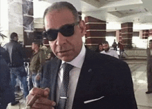 a man wearing sunglasses and a suit is smoking a cigar .
