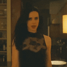 a woman in a black lace top is standing in a kitchen looking at the camera .