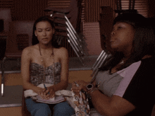 two women sit in front of a stack of chairs one is writing in a notebook