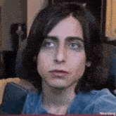 a young man with long hair and blue eyes is sitting in a chair .
