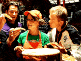a woman in an apron is holding a pizza and smiling while two men eat pizza