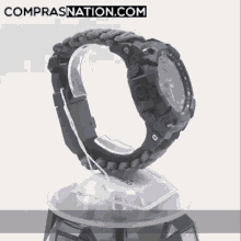 a watch with a paracord band sits on a display stand with the website comprasnation.com written on the bottom