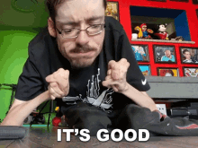 a man with glasses is sitting on the floor with his hands on his knees and the words `` it 's good '' above him .