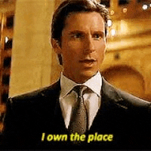a man in a suit and tie is standing in front of a building and says `` i own the place '' .