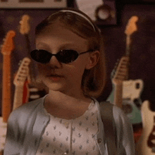 a young girl wearing sunglasses is standing in front of a wall of guitars