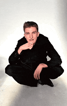 a man in a black jacket is squatting down with his hand on his chin