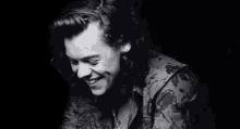 harry styles is smiling in a black and white photo . he is wearing a floral shirt and a necklace .
