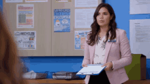 a woman in a pink jacket holds a clipboard in front of a bulletin board that says saving lives