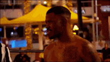 a man without a shirt is standing in a boxing ring with a yellow tent in the background .