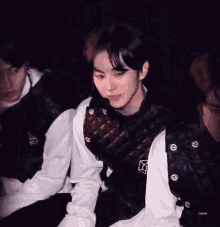 a group of young men are sitting in a dark room and one of them is wearing a black vest with a check logo on it