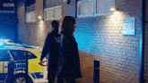 a man and a woman are walking in front of a police car that says bbc on it