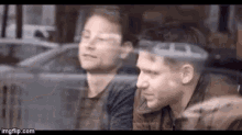 two men are sitting next to each other in front of a window and looking out .