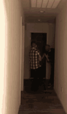 a man in a plaid shirt stands next to another man in a black shirt in a hallway