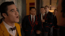 a man in a yellow suit and bow tie is talking to another man in a black suit