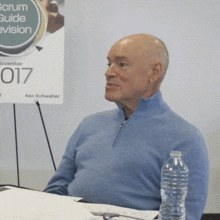 a man sitting at a table with a bottle of water in front of a sign that says scrum guide revision