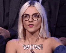 a woman wearing glasses and a ring is sitting in front of a crowd and says wow .