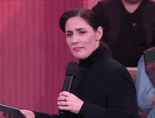 a woman in a black turtleneck is holding a microphone and looking at the camera