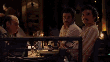 a man with a mustache sits at a table with other men in a dark room