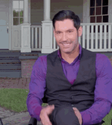 a man in a purple shirt and black vest smiles while sitting in front of a house