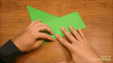 a person folding a piece of green paper on a wooden table with the words made in animatica on the bottom