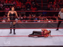 a woman is laying on the ground in a wrestling ring while a referee stands behind her .