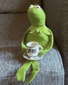 kermit the frog is sitting on a couch holding a cup of coffee and saucer .