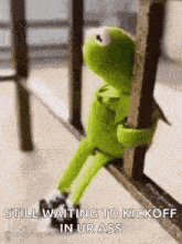 kermit the frog is sitting on a railing holding onto a railing and waiting to kick off in ur ass .