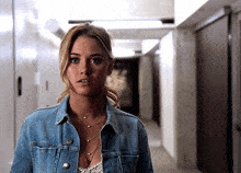 a woman wearing a denim jacket and a necklace is standing in a hallway