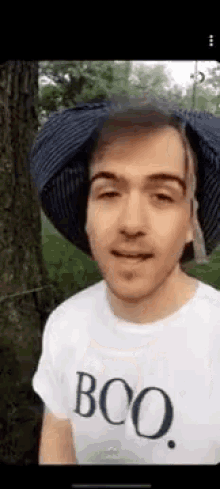 a man wearing a hat and a t-shirt with the word boo on it is standing next to a tree .