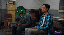 a man in a plaid shirt sits next to another man with a crocodile on his face