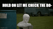 a statue of a man 's head is sitting in the grass next to a blue barrel and a truck .
