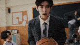 a man in a suit and tie is holding a basketball in a classroom with other students .