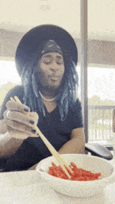 a man with blue dreadlocks is eating a bowl of macaroni and cheese with chopsticks