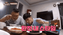 two men are sitting at a table with plates of food and a foreign language written on the table