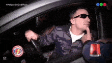 a man wearing sunglasses is driving a car with a peligroso sin codificar logo behind him
