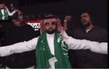 a man with a green scarf around his neck is surrounded by other men with their arms outstretched .