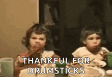 two little girls are sitting at a table eating food and the words thankful for drumsticks are on the screen .