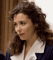 a woman with curly hair is wearing a blue jacket with a white collar and a badge with the letter t on it