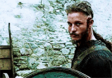a man with a beard and long hair is holding a shield and looking at the camera .